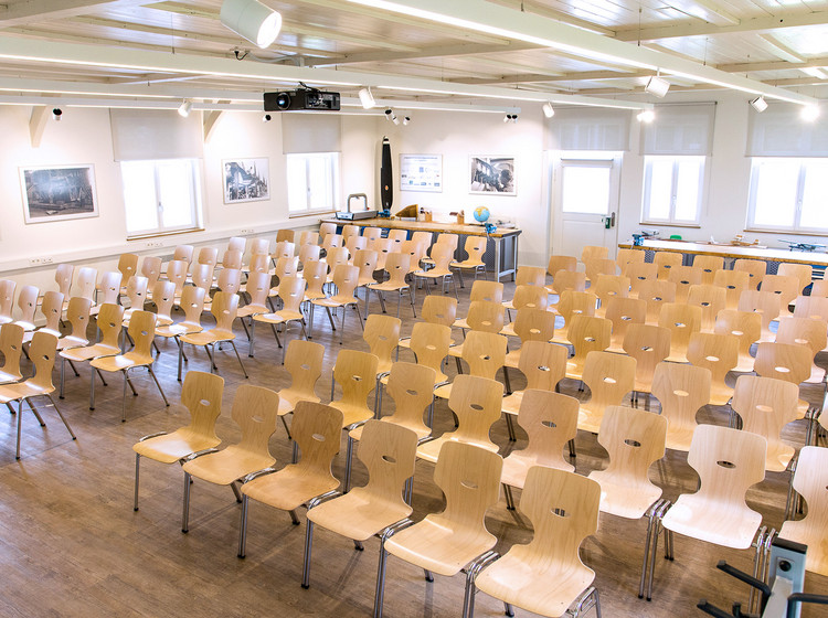 Seminarraum mit zahlreichen Holzstühlen in Reihenanordnung, einige mit Schreibflächen ausgestattet, vorne eine Leinwand und Rednerpult.