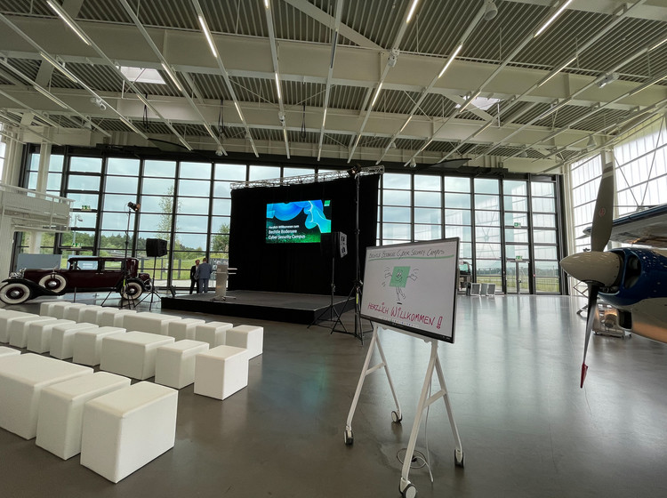 Veranstaltungsbereich mit weißen Sitzbänken, großer Präsentationsleinwand und Flipchart, umgeben von historischen Flugzeugen in heller Halle mit Glasfassade.