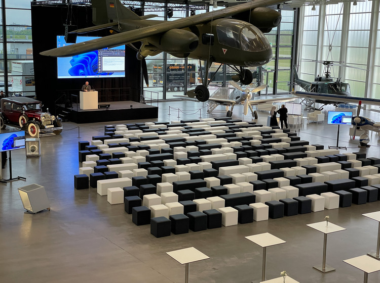 Moderner Veranstaltungsraum mit schwarz-weißen Sitzwürfeln vor einer Bühne mit Präsentation, im Hintergrund große Fensterfront und hängendes Flugzeug.