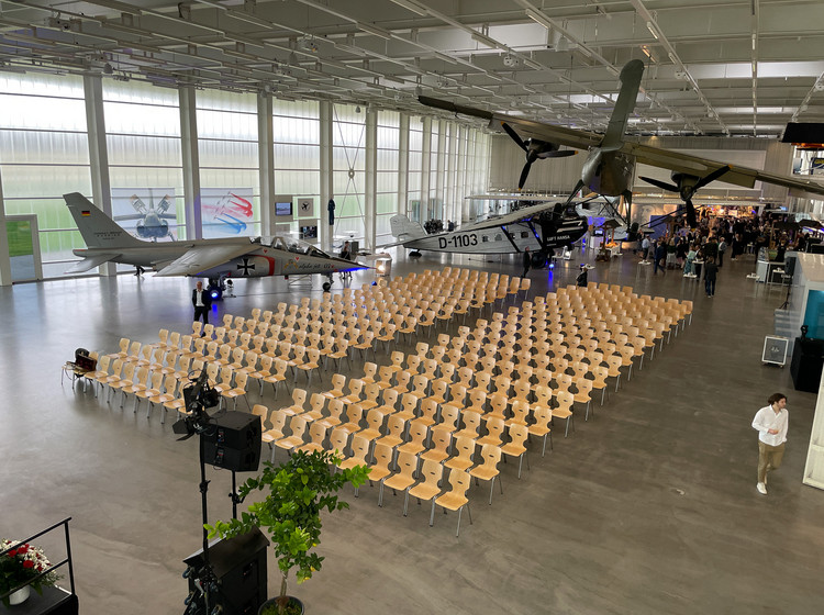 Großer Veranstaltungsraum mit zahlreichen Holzstühlen in Reihen, flankiert von historischen Flugzeugen und Hubschraubern in einer modernen, lichtdurchfluteten Halle.