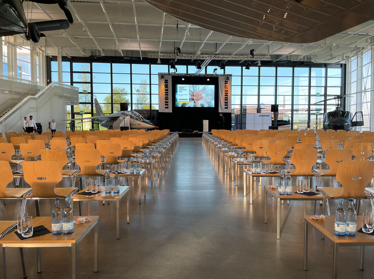 Großer Veranstaltungsraum mit Holzstühlen in Reihen, auf jedem Platz ein Headset, Bühne mit Leinwand im Hintergrund, Glasfassade und Flugzeuge sichtbar.