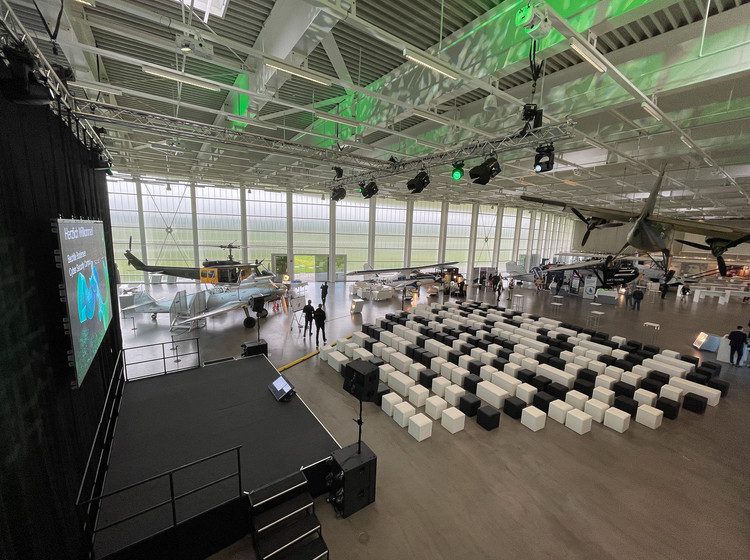 Großer Veranstaltungsbereich mit schwarzer Bühne und Leinwand, lockerer Bestuhlung aus schwarzen und weißen Würfeln, grün beleuchteter Decke und historischen Flugzeugen in heller Halle.