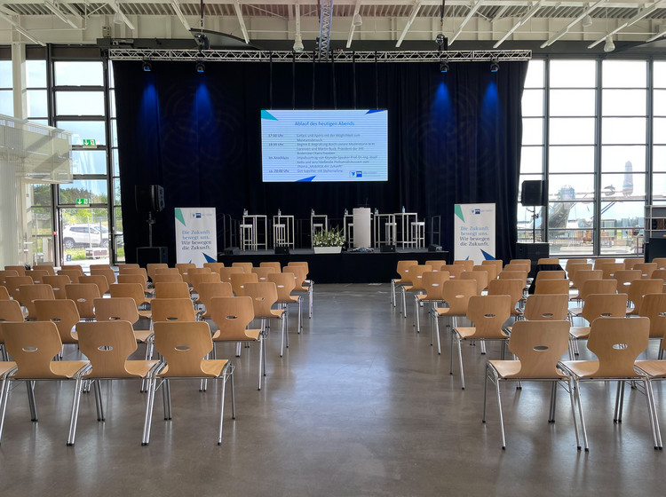 Veranstaltungssaal mit Reihen aus Holzstühlen, ausgerichtet auf eine Bühne mit Podium, großen Displays und blauer Beleuchtung, Glasfassade im Hintergrund.
