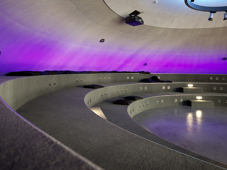 Futuristisch gestalteter Ausstellungsraum mit geschwungenen Wegen, indirekter Beleuchtung und violett angestrahlter Wand unter einer gewölbten Decke.